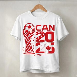 May include: A white short-sleeve t-shirt featuring a red graphic design of a soccer ball atop a trophy shape, with the text CAN 2026 and a small maple leaf icon integrated into the number 6, all printed in a bold red color on the front.