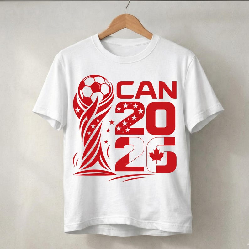 May include: A white short-sleeve t-shirt featuring a red graphic design of a soccer ball atop a trophy shape, with the text CAN 2026 and a small maple leaf icon integrated into the number 6, all printed in a bold red color on the front.