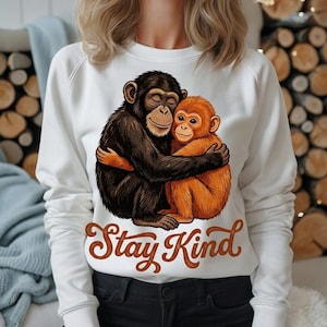 Pode incluir: Camisola branca com um gráfico de dois macacos abraçados, um preto e outro laranja. O texto "Stay Kind" está escrito em castanho por baixo. A camisola tem mangas compridas e gola redonda.