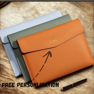 Könnte beinhalten: Drei Laptop-Hüllen aus Leder in Hellblau, Grün und Orange. Die orangefarbene Hülle ist mit dem Namen "Lisa" in Gold geprägt. Ein schwarzer Stift liegt auf der Holzoberfläche. Der Satz "FREE PERSONLISATION" ist unten aufgedruckt.
