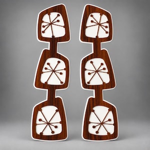 May include: Two vertical wall art panels featuring a dark brown wood grain finish with white cutouts. Each panel consists of three stacked, irregular geometric shapes, each containing a brown starburst pattern with thin lines and circular ends.