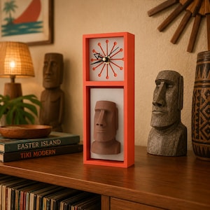 May include: A rectangular orange clock with a white face featuring a sunburst design and black hands. Below the clock face, a three-dimensional brown Moai statue head is mounted on a white rectangular panel within the orange frame.
