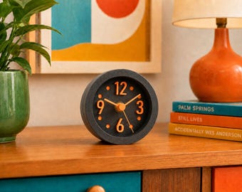 May include: A round analog desk clock with a dark gray textured frame and a black face. The clock features bright orange raised numerals at the 12, 3, 6, and 9 positions, with orange dots for the other hours and matching orange clock hands.