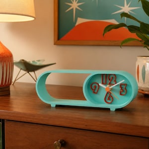May include: A horizontal, oval-shaped turquoise desk clock with a hollow left side and a solid right side containing the clock face. The clock face features raised, dark orange numerals and white hands, with a small orange center cap.