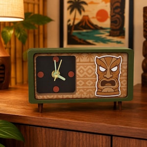 May include: A rectangular olive green desk clock with four small wooden legs. The clock face features a black square with red circular markers and light green hands. The right side displays a tan background with a brown tiki mask graphic.