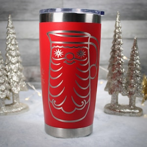 May include: A red insulated tumbler with a silver lid and base. The tumbler features a silver etched design of a pitcher with star and wave details. The design is centered on the tumbler. The background includes decorative silver Christmas trees.