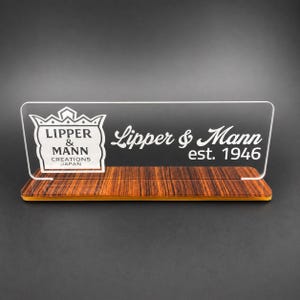 May include: A clear rectangular acrylic sign featuring the white printed text Lipper & Mann Creations Japan and Lipper & Mann est. 1946. The sign is mounted on a rectangular base with a dark brown wood grain pattern and a visible orange edge.