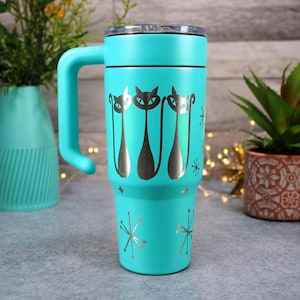 May include: A bright turquoise stainless steel tumbler with a handle and a clear lid. The exterior features a metallic silver graphic of three stylized cats standing side by side, accented with scattered starburst and diamond-shaped patterns.