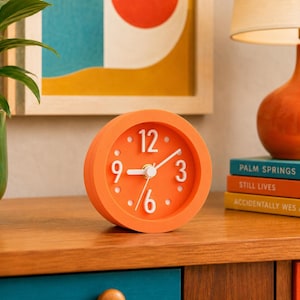 May include: A round, bright orange analog alarm clock with a matching orange face, white hour markers, and white numerals at the 3, 6, 9, and 12 o'clock positions. The clock features white hour, minute, and second hands centered on the dial.