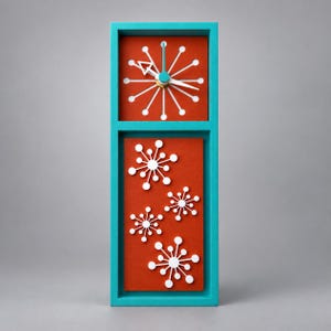May include: A rectangular wall clock with a bright teal frame and a deep orange face. The upper section features a white starburst clock dial with white hands. The lower section displays four white starburst decorative elements on the orange background.