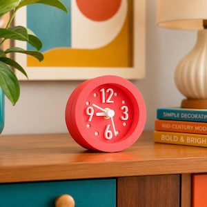 May include: A round, bright red analog clock with a matte finish, featuring white raised numerals for 3, 6, 9, and 12, along with white hour and minute hands and a central gold-toned pin. The clock face is a matching red with white dot markers.