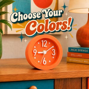 May include: A round orange analog alarm clock with white numbers and hands. The clock face features the numbers 3, 6, 9, and 12 in a bold font. Above the clock, the text Choose Your Colors is displayed in a stylized, multi-colored font.