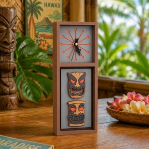 May include: A rectangular brown clock with a sunburst-style face featuring orange spokes and black hands. Below the clock face, two stacked tiki-style masks with dark gray faces, tan borders, and glowing red eyes are mounted on a gray background.