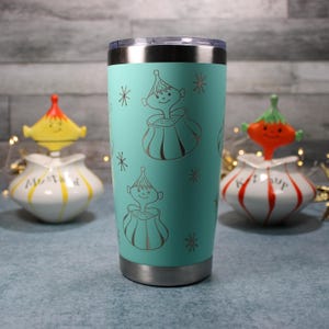 Pode incluir: Um copo térmico turquesa com aro e base prateados, apresentando um design gravado repetido de um personagem caprichoso em um pote com flocos de neve. Potes decorativos de mostarda e ketchup ao fundo.