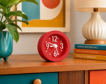 May include: A round, red analog clock featuring a matte finish, white raised numerals at the three, six, nine, and twelve o'clock positions, and white hour and minute hands with a central gold-toned pin, designed for tabletop placement.