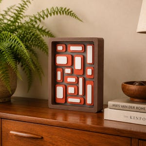 May include: A rectangular wall art piece featuring a dark brown frame containing a gray background with a layered arrangement of orange and white rounded rectangular shapes of varying sizes, creating a three-dimensional geometric composition.