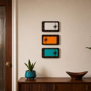 May include: Three rectangular wall plaques with dark wood frames, each featuring a starburst design. The top plaque has a white center, the middle plaque has an orange center, and the bottom plaque has a teal center, all arranged vertically.