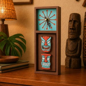 May include: A rectangular brown wooden clock featuring a light blue clock face with a red sunburst design and black hands. Below the clock face, two stacked, carved tiki-style masks in red and light blue are mounted on a brown background.