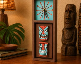 May include: A rectangular brown wooden clock featuring a light blue clock face with a red sunburst design and black hands. Below the clock face, two stacked, carved tiki-style masks in red and light blue are mounted on a brown background.