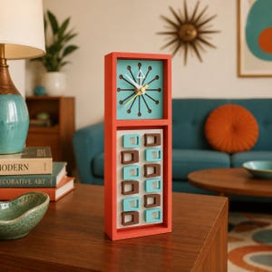 May include: A tall, rectangular orange clock featuring a teal face with a black starburst design and white hands. The lower section displays a vertical grid of alternating teal and brown rectangular cutouts on a white background.