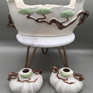 May include: A white ceramic planter with a wavy rim, brown handles, and green and brown tree designs. Two matching small vases are also present. The planter sits on a white marble-topped stand with gold legs. Decorative pottery.