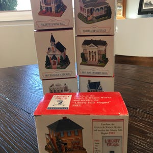 May include: A collection of miniature building figurines in boxes. The boxes feature images of a church, a bank, a cottage, and a music hall. The boxes are labeled with the names of the buildings and the "Liberty Falls Collection" logo.