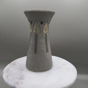 May include: A gray ceramic vase with a textured surface and a flared top. The vase features gold-colored accents and a dark interior. The vase is displayed on a white marble-topped side table with gold legs.