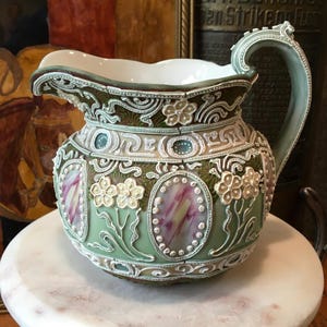 May include: A sage green ceramic pitcher featuring intricate white relief scrollwork, raised floral motifs, and oval panels with a marbled pink and white finish. The vessel has a wide spout, a curved handle, and a smooth white interior surface.