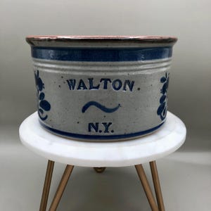May include: A round, stoneware crock with a blue and gray speckled finish. The crock features the word "WALTON" and "N.Y." in blue lettering, along with a wavy line and floral designs. The rim is a reddish-brown color.