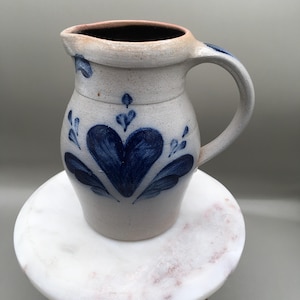 May include: A light gray stoneware pitcher featuring a hand-painted dark blue heart motif on the front. The ceramic vessel has a curved handle, a wide spout, and a speckled texture throughout the body, showcasing a classic salt-glaze style finish.