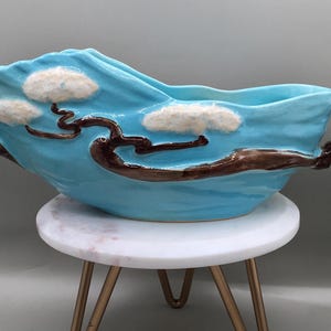 May include: A turquoise ceramic bowl with a wave-like design, featuring brown branch-like handles and white cloud accents. The bowl is displayed on a small, white marble-topped table with gold legs. A decorative piece.
