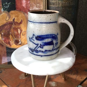 May include: A speckled light gray ceramic mug featuring a dark blue embossed pig graphic on the front. The mug has a sturdy handle, a cylindrical body with horizontal blue bands, and a dark interior. The surface has a textured, rustic appearance.