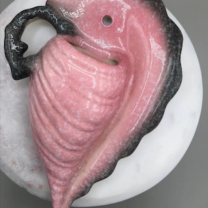 May include: A pink ceramic leaf-shaped dish with a black handle and rim. The dish has a hole near the top and a textured surface. The dish is on a white marble surface.