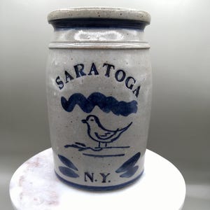 May include: A gray stoneware jar with a blue rim and base. The jar features the word "SARATOGA" in blue, along with a blue wave design and a bird illustration. The text "N.Y." is at the bottom.