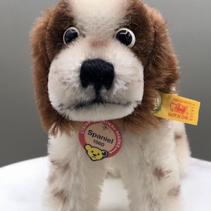 May include: A plush Spaniel dog toy with brown and white fur, black eyes, and a black nose. The toy has a red and white tag that reads "Spaniel 1960" and a yellow tag that reads "Knopf im Ohr".