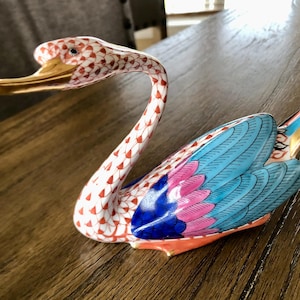 May include: A decorative ceramic swan figurine. The swan features a white body with a red and white pattern on the neck. The wings are blue and pink with gold accents. The beak is gold.
