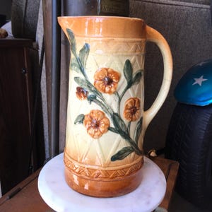 May include: A ceramic pitcher featuring a cream and orange gradient glaze with a raised, embossed diamond pattern around the top and bottom. The center is decorated with a relief design of orange flowers and green leaves on a textured surface.