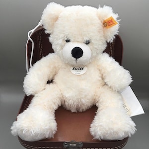 May include: A cream-colored Steiff teddy bear with a black nose and eyes, sitting in a brown suitcase. The bear has a fluffy texture and a tag on its ear. The Steiff logo is visible on the bear's chest.