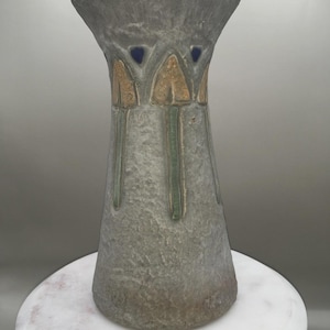 May include: A gray ceramic vase with a textured surface. The vase has a flared top and a tapered body. It features gold and green accents, with small blue details near the top. The vase is displayed on a white marble surface.