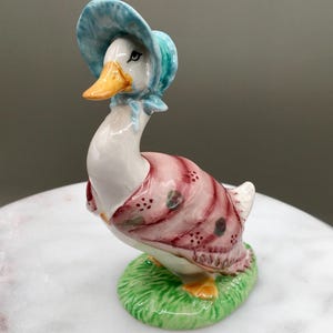 May include: A ceramic duck figurine wearing a blue bonnet and a pink and red striped dress. The duck has an orange beak and feet, and stands on a green grassy base. The figurine is a decorative item.