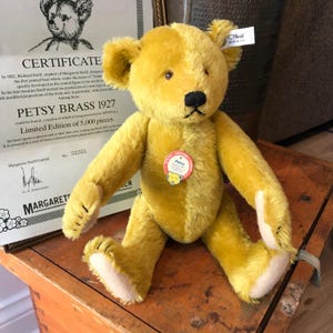 May include: A golden-yellow mohair teddy bear with a black stitched nose and mouth. The bear features a white ear tag labeled Steiff and a round red and white chest tag that reads Petsy Replica 1927. It has light-colored felt paw pads.