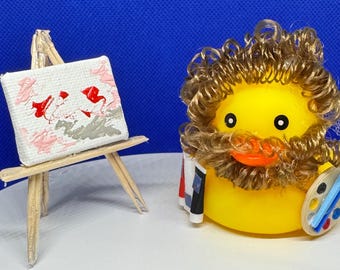 May include: A yellow rubber duck wearing a brown wig and holding a paintbrush and palette. Beside the duck is a miniature wooden easel with a small canvas displaying a painting with red, pink, and gray brushstrokes. The background is a solid blue.