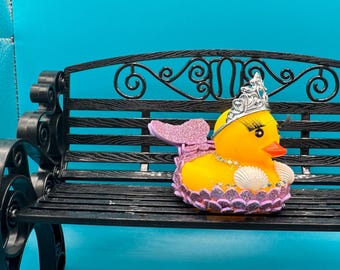 May include: A yellow rubber ducky with a silver tiara, long eyelashes, and a seashell necklace. The duck has a pink glittery mermaid tail and a matching collar. It is sitting on a black metal bench.