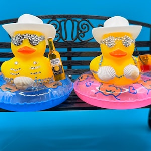 May include: Two yellow rubber ducks wearing sunglasses and white hats, resting on inflatable rings. One duck holds a bottle of Corona Extra. The ducks are decorated with rhinestones and seashells, sitting on a black bench.