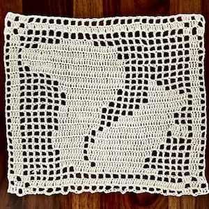 Op de afbeelding: Crèmekleurige, vierkante gehaakte doily met een bloemenontwerp in het midden. De doily is gemaakt met een strak steekpatroon en heeft een geschulpte rand. De doily is waarschijnlijk handgemaakt en geschikt voor woondecoratie.