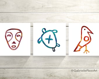 May include: Three watercolor paintings on white canvases, each featuring a different symbol. The first painting shows a stylized human face in shades of purple and brown. The second painting shows a stylized turtle in shades of blue and green. The third painting shows a stylized bird in shades of orange and red. The paintings are arranged in a row on a white wooden background. The text "@GabrielleRicciArt" is visible at the bottom of the image.