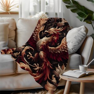 May include: A decorative throw blanket featuring a phoenix design in shades of red, gold, and brown against a black background. The blanket is draped over a white sofa, showcasing the intricate details of the artwork. The blanket adds a touch of elegance to any living space.