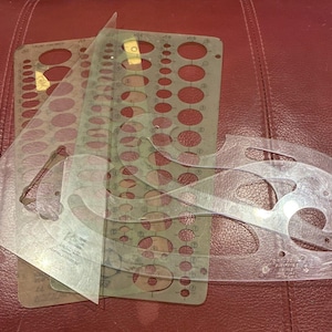 May include: A collection of drafting tools, including clear plastic French curves, a triangle ruler, and a stencil with various circle sizes. The tools are arranged on a reddish-brown surface, suggesting a workspace.