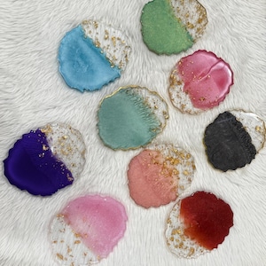 May include: A collection of handmade resin coasters in various colors, including blue, green, pink, and purple. Each coaster features a gold leaf design and a clear, textured edge. The coasters are round and sit on a white, fluffy surface.