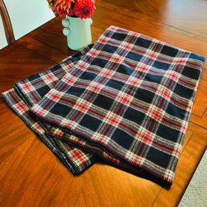 May include: A folded rectangular blanket featuring a dark navy blue, white, and bright red plaid pattern. The fabric has a soft, textured appearance with visible stitching along the edges, creating a classic grid design across the entire surface.